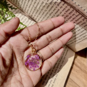 A delicate pendant with a real purple flower preserved in a clear resin oval, hanging from a thin, gold-plated sterling silver chain.